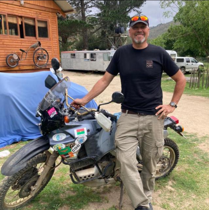A picture of Kevin Bake the owner and founder of Black Mountain Adventures standing next to a motorcycle with a wooden cabin and beautiful green tress in the background